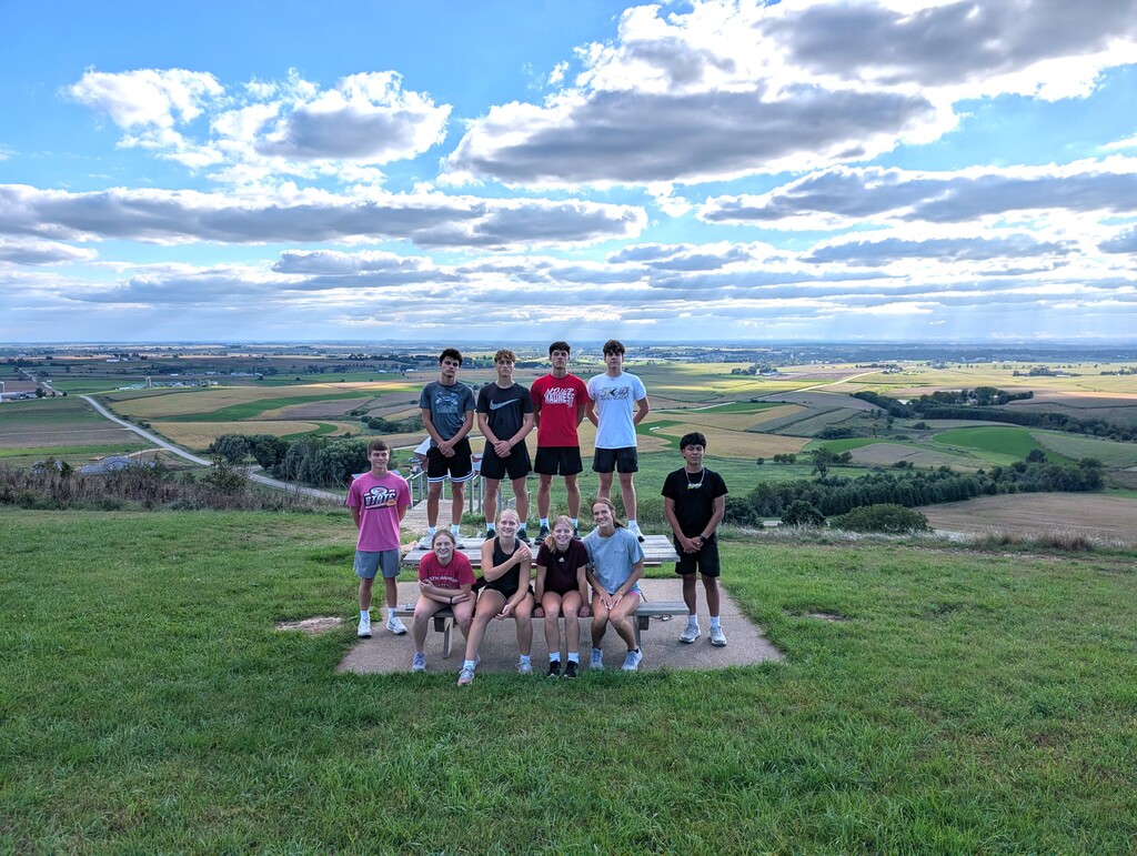 Group of people in a grassy field. Some stand on concrete, others sit. Background shows hills and sky.