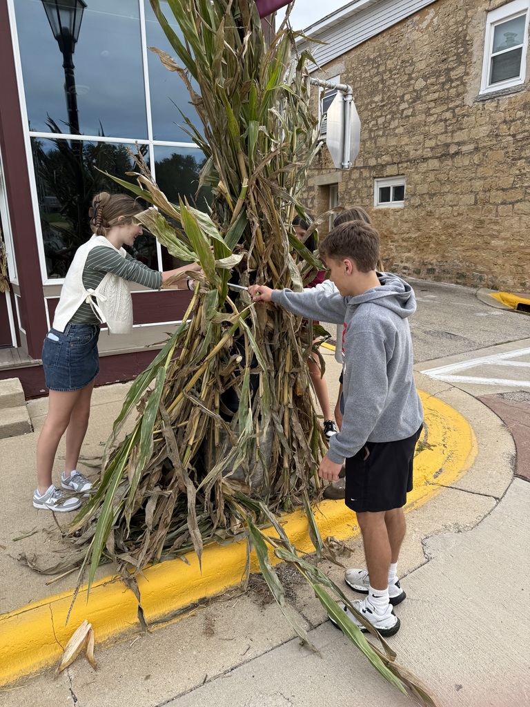 Three people work together to assemble a tall corn stalk decoration on a city sidewalk.