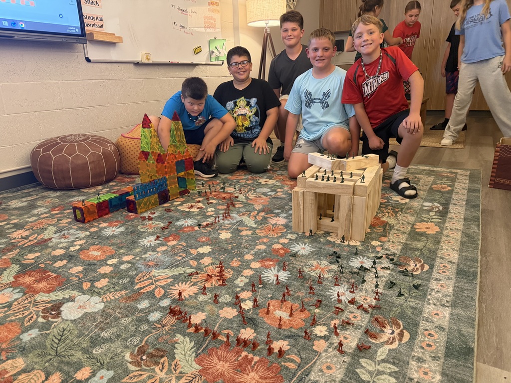 A group of children is smiling and posing for a photo, surrounded by wooden blocks and a patterned rug.