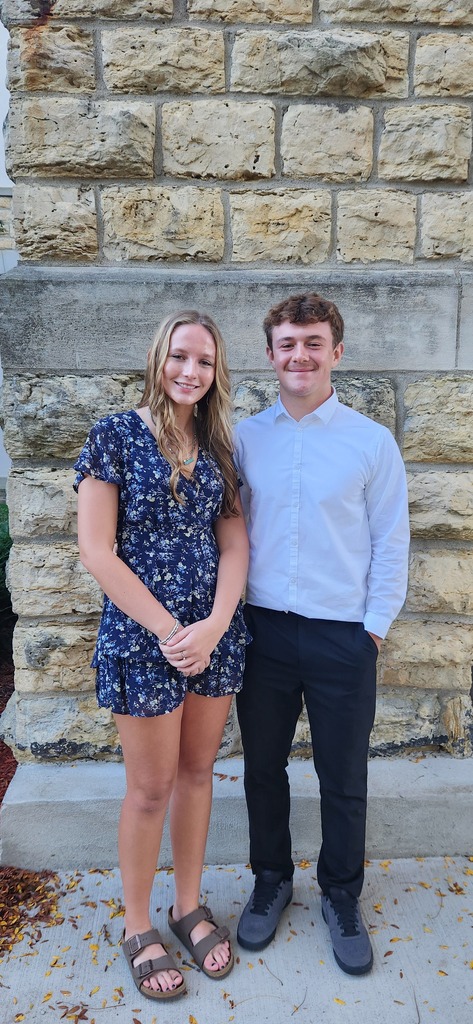 A man and woman stand together outside in front of a textured stone wall.
