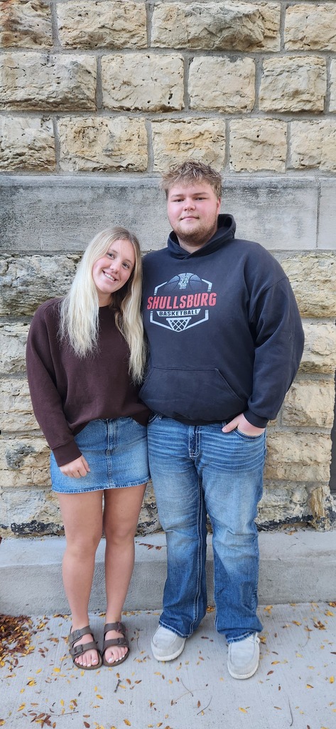 Couple standing together in front of a stone wall, wearing casual clothes; one in a brown shirt, denim skirt, and sandals, the other in a blue hoodie and jeans.