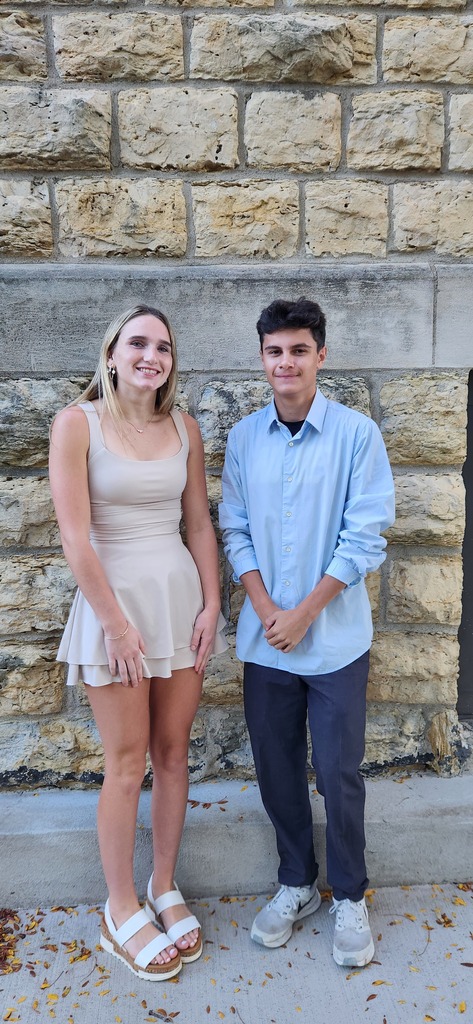 A woman and a man stand together outside near a brick wall. The woman wears a dress and sandals, and the man wears a shirt and pants.