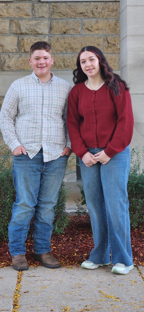 Two people, one in white and one in red, standing outside a building with stone walls.