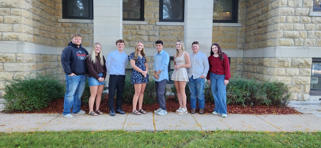 Seven people pose for a photo in front of a brick building with large windows.