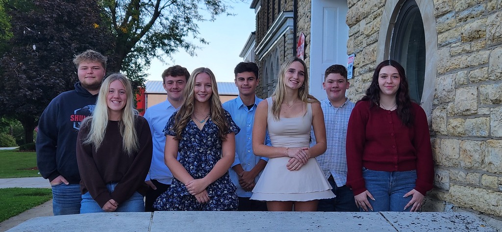 A group of young adults stands together in front of a building with trees and grass.