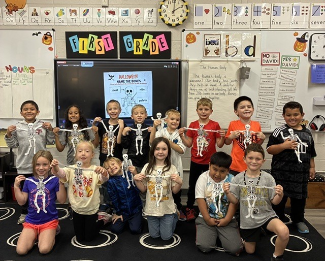 Group of students in a classroom, wearing skeleton outfits and posing for a photo. Whiteboard behind them with "First Grade" text.