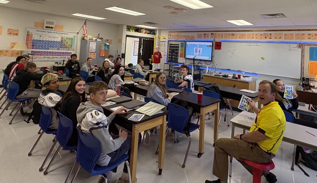 Students in a classroom with desks and chairs, an instructor in front, and a screen on the wall.