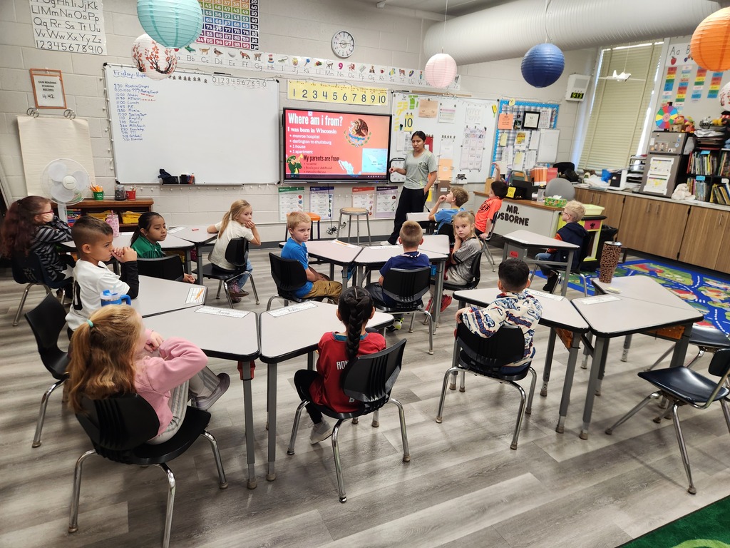 Classroom with students seated at desks, a teacher at a front screen, and a whiteboard and balloons in the background.