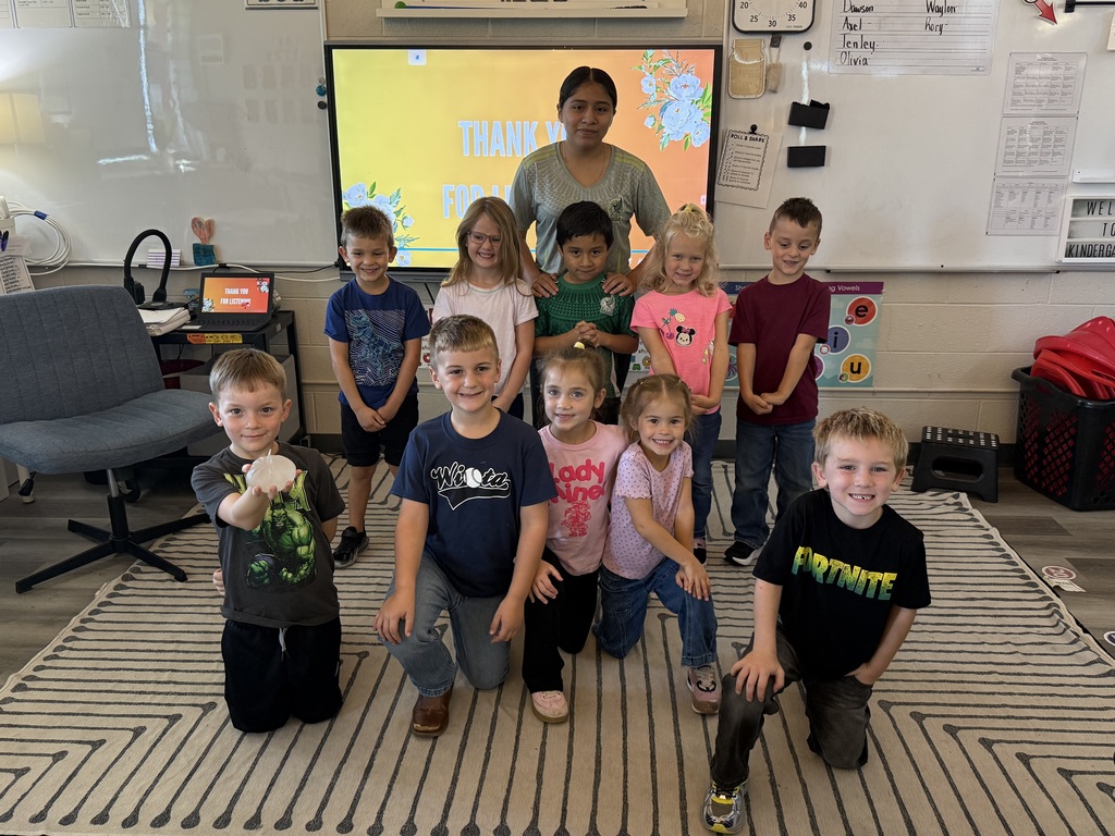 Several children pose for a photo in a classroom with a woman standing behind a whiteboard and a television screen.