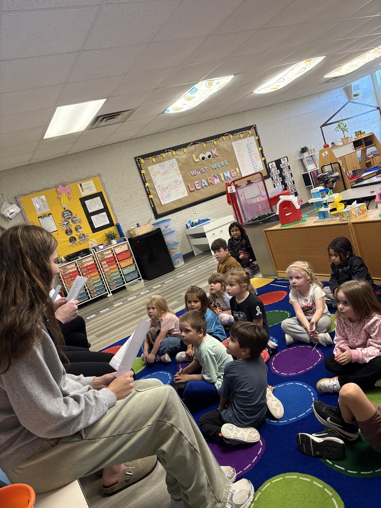 Woman reading to children on a colorful rug in a classroom with bulletin boards and furniture.