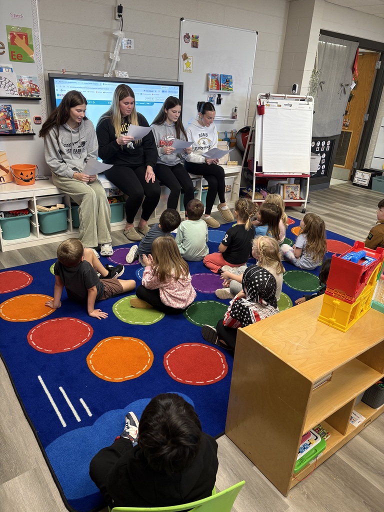 Group of young children sitting in circles on a rug with teachers. Colorful rug and shelves in background.