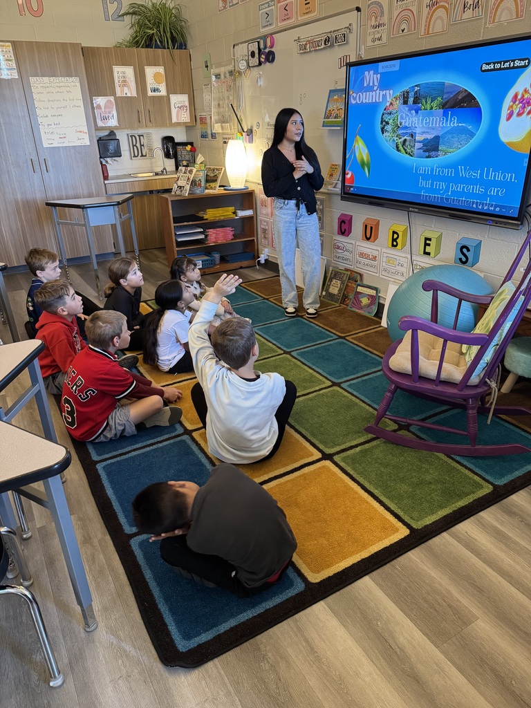 Children sit on a colorful rug, attentively listening to a teacher standing in front of a large screen.