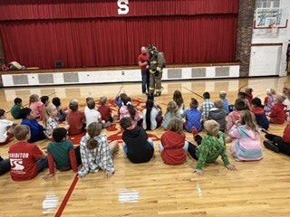 A classroom with children in a circle on a basketball court, a man in a red uniform standing.
