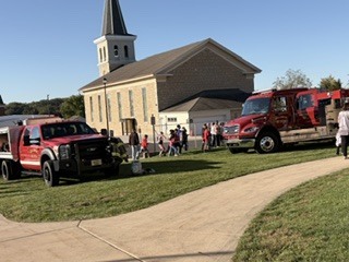Two red fire trucks parked on a lawn in front of a church. Several people stand around.