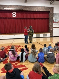 A firefighter in uniform stands with a woman before a seated classroom of students. Behind them, a red curtain hangs over a stage.