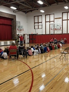Inside a gym, a group of children sit on the floor, and a person stands near the basketball hoops.