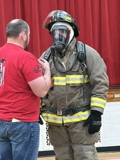 A man in a red shirt stands next to a firefighter in full gear, including a mask and gloves.