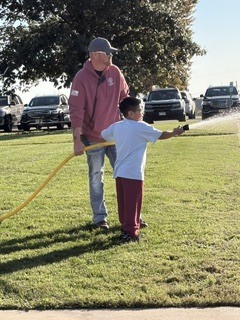 A man and a child water a grassy area with a hose, near parked cars.