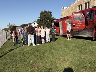 A group of people stand in a circle around a red fire truck. They are on a grassy field with a building and trees in the background.