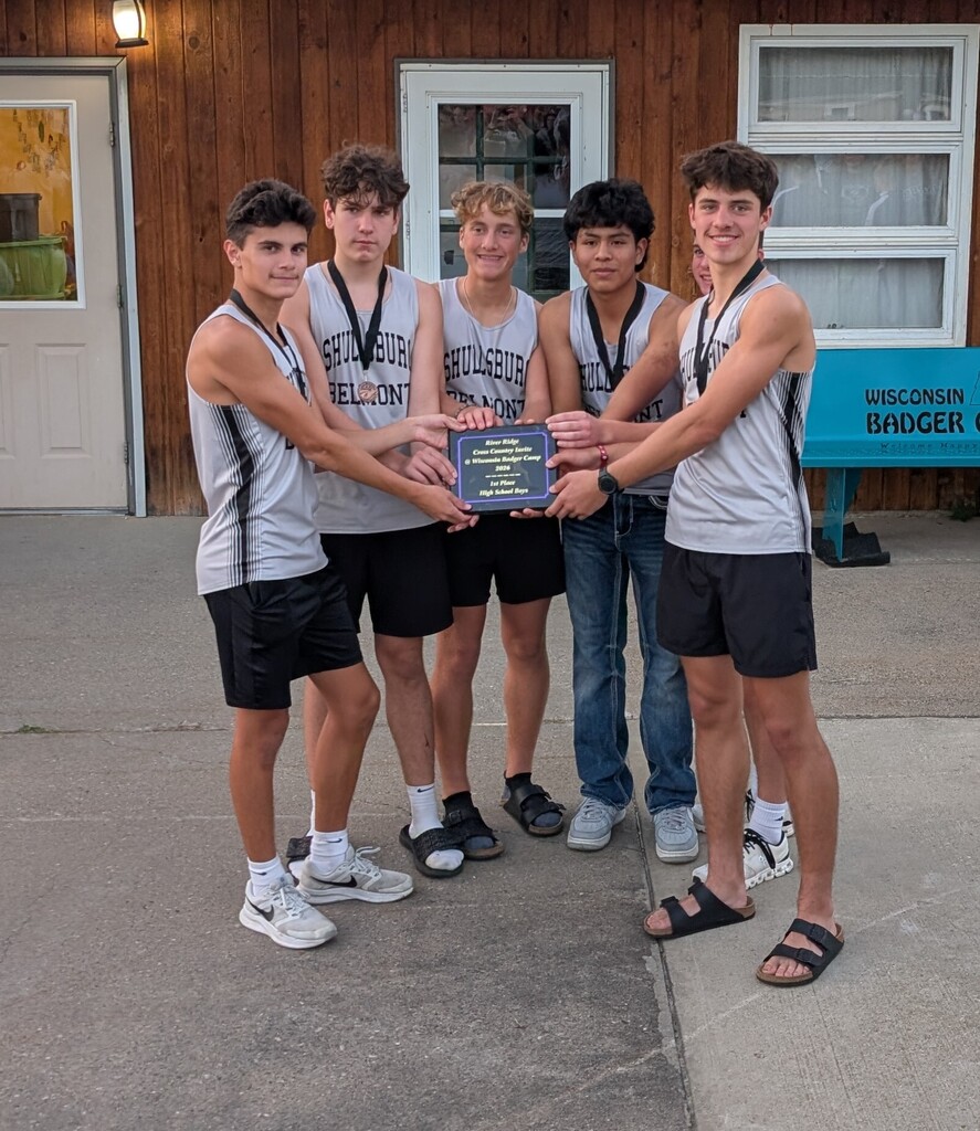 Six boys in athletic wear pose for a photo holding a plaque in front of a building.