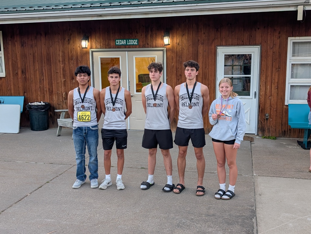 Five young athletes, three males and two females, stand together, smiling, with medals around their necks. Behind them is a building.