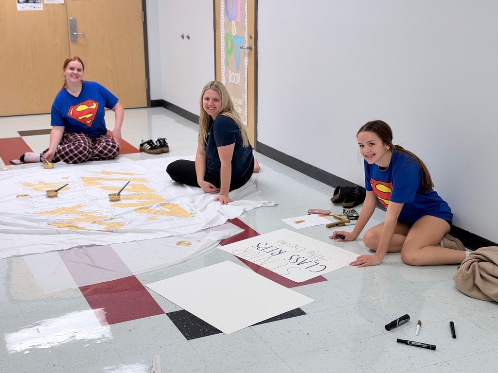 Three people sit on the floor, one holding a marker, with a large white cloth and papers on the floor.