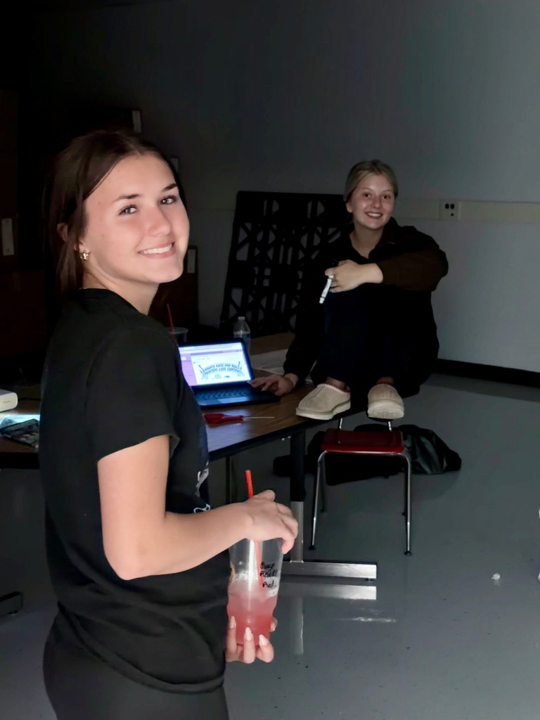 Two women, one seated with a laptop and one standing holding a drink, are in a room with a table.