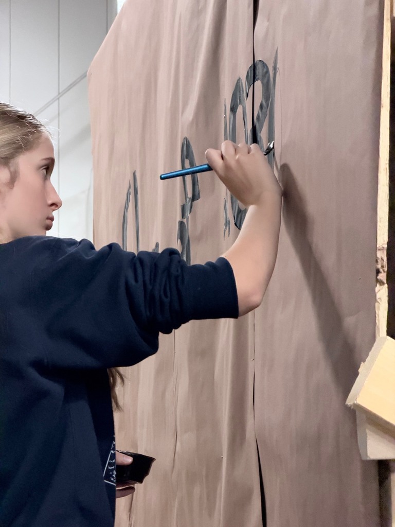 A person in a blue shirt painting black writing on a brown fabric. A light overhead illuminates the scene.