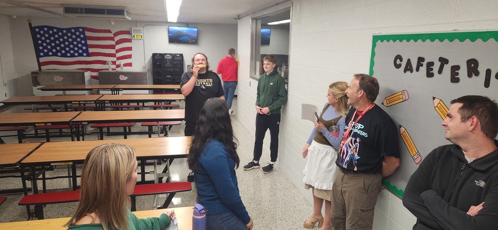 A group of people in a classroom. An American flag is displayed. A wall with the word "cafeteria" is visible.