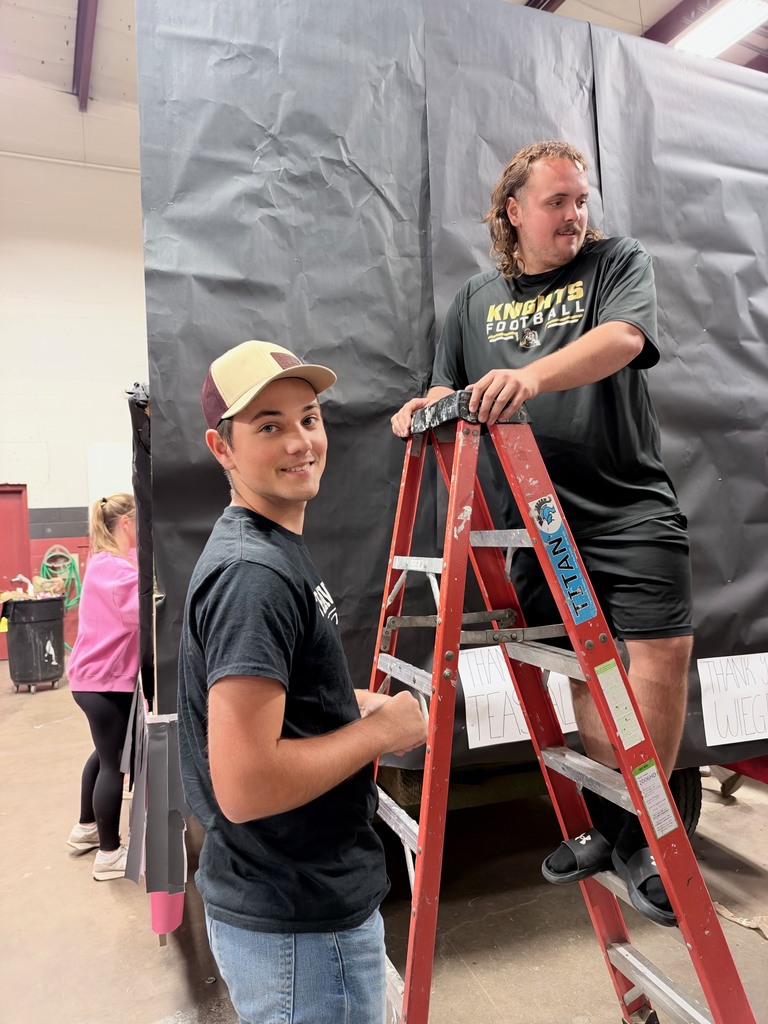 Two people are working on a tall ladder, one standing and one seated. They wear casual clothing. A large, black fabric is behind them.
