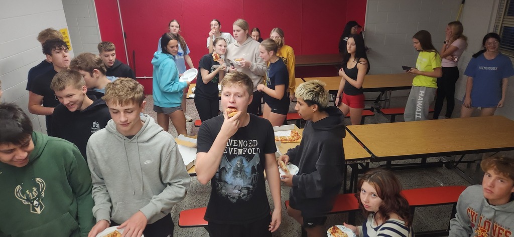 A group of students gathered in a cafeteria, some eating food, with tables and red wall in background.