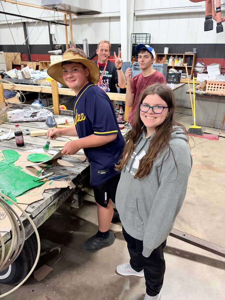 Four people, two adults and two children, stand around a work table in a workshop. One child is wearing a cowboy hat.