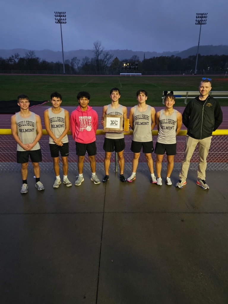 Six young men stand on a track, smiling and holding a trophy. They wear matching gray shirts and black shorts. A man stands behind them wearing a black jacket and khaki pants.