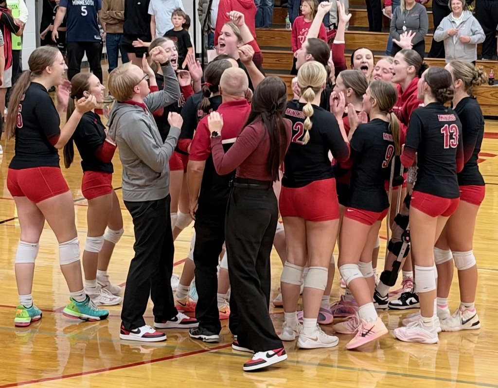 Group of young women in matching black uniforms and red shorts huddle together on a court.
