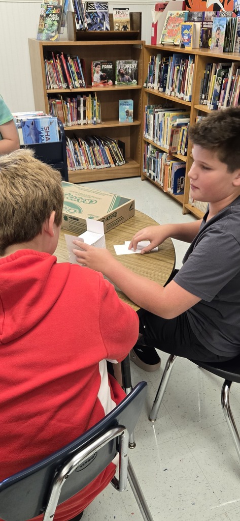 Two boys seated in library, one holds paper, the other holds a box, a table between them.