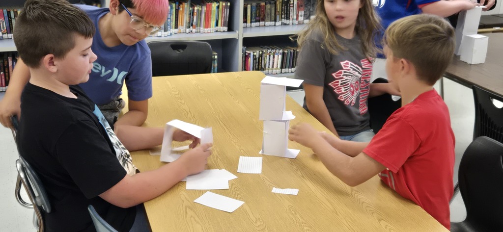 Four children sitting around a table in a library, constructing and examining a structure made of paper.