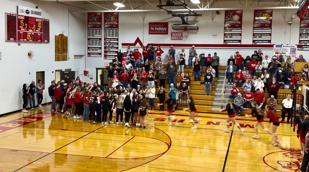 Many people stand and sit on bleachers in a gym.
