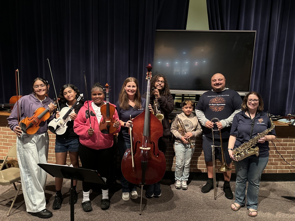 District staff members and students are posing with a variety of musical instruments in a school auditorium.