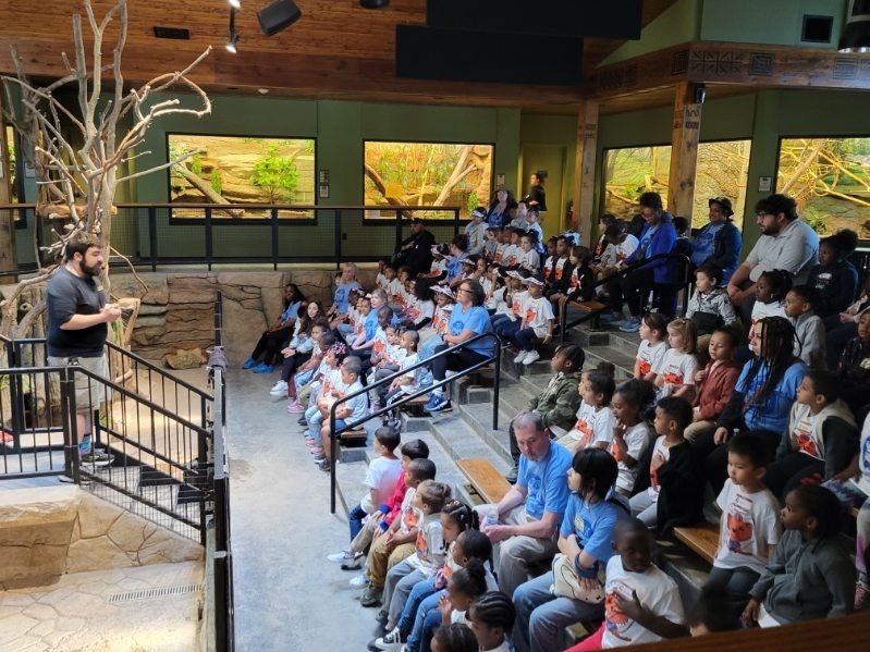 A speaker standing on a stage in front of a seated audience of children and adults at a venue, with trees in the background and large windows showing greenery outside.