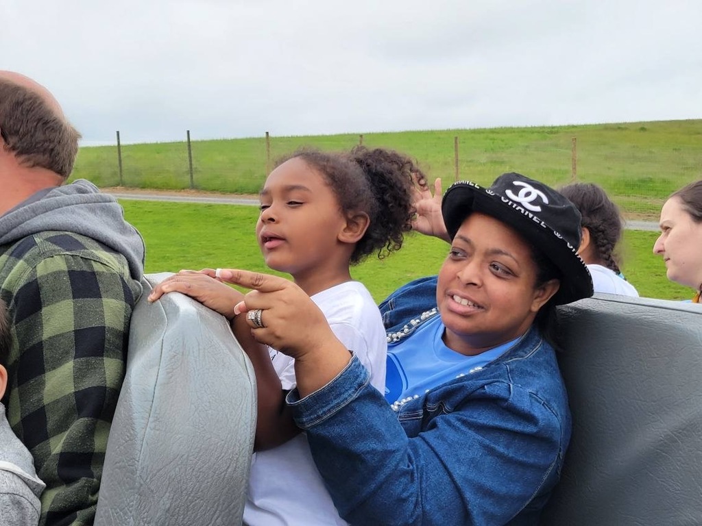Superintendent of Schools, Dr. Berry-Brown wearing a black hat with a young student sitting on her lap, both engaged in attentive expressions during a drive-thru safari. Surrounded by other passengers, they are outdoors with a grassy landscape in the background.