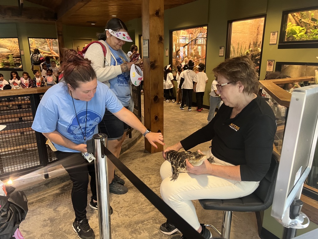 A woman in a blue shirt points at a turtle held by another woman sitting on a chair. In the background, a group of children in matching shirts observe the interaction. The setting appears to be an educational center with animal exhibits.
