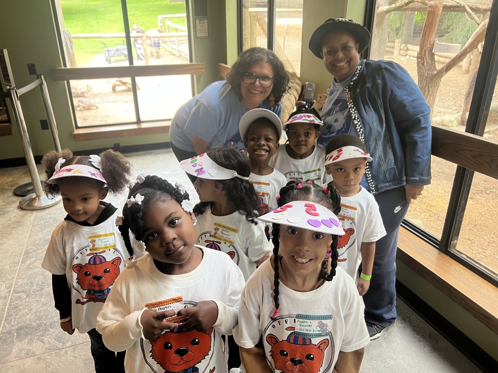 A group of students wearing matching t-shirts with a cartoon lion and visors pose for a photo indoors during a day at the zoo. Superintendent of Schools, Dr. Berry-Brown and a Pre-K teacher are both smiling. A green outdoor area is visible through a window.