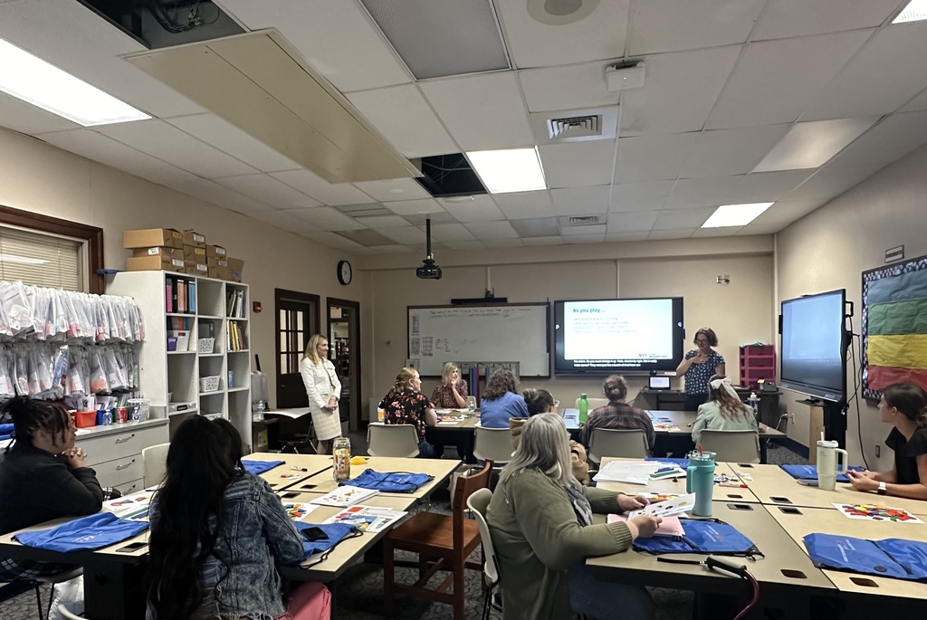 A classroom filled with attendees facing a presentation on monitors, engaged in discussions and activities at tables with supplies.