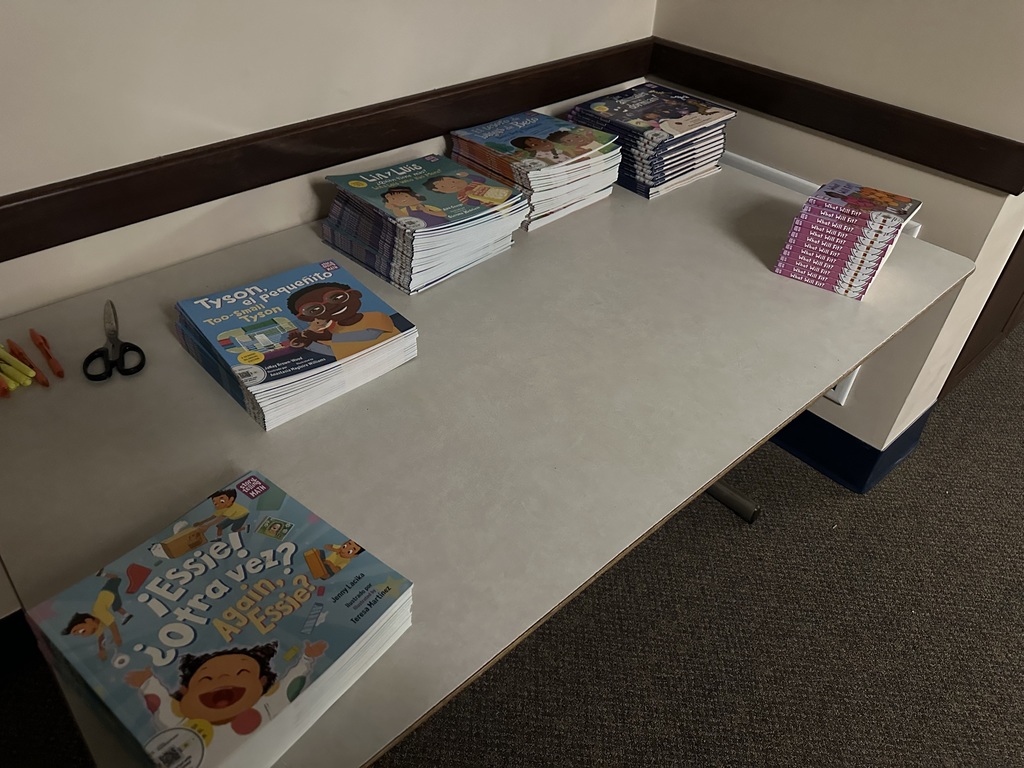 A table displaying stacks of colorful children's books, with scissors and highlighters nearby, in a well-lit room.