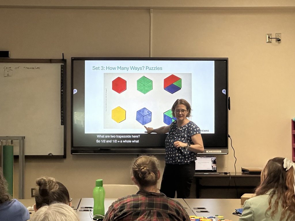A woman presents geometric puzzles on a screen to an engaged audience in a classroom, with manipulatives spread across tables.
