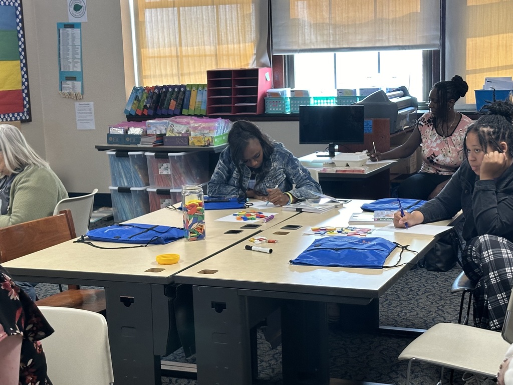 A classroom setting with adults engaged in crafts and writing, colorful supplies scattered on tables, and shelves filled with materials.
