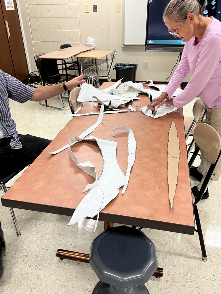 Two people are working at a table covered with fabric patterns and cutouts, engaged in a crafting or sewing project in a classroom.
