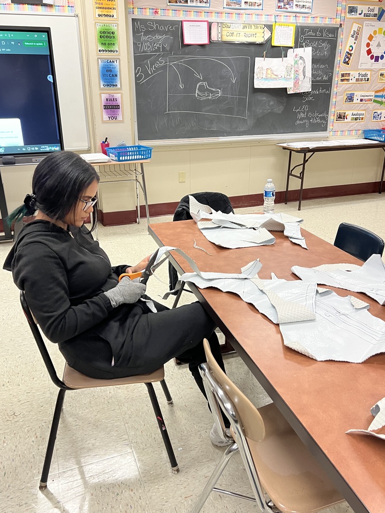 A student wearing gloves sits at a table, cutting fabric pieces, with a classroom and chalkboard in the background.