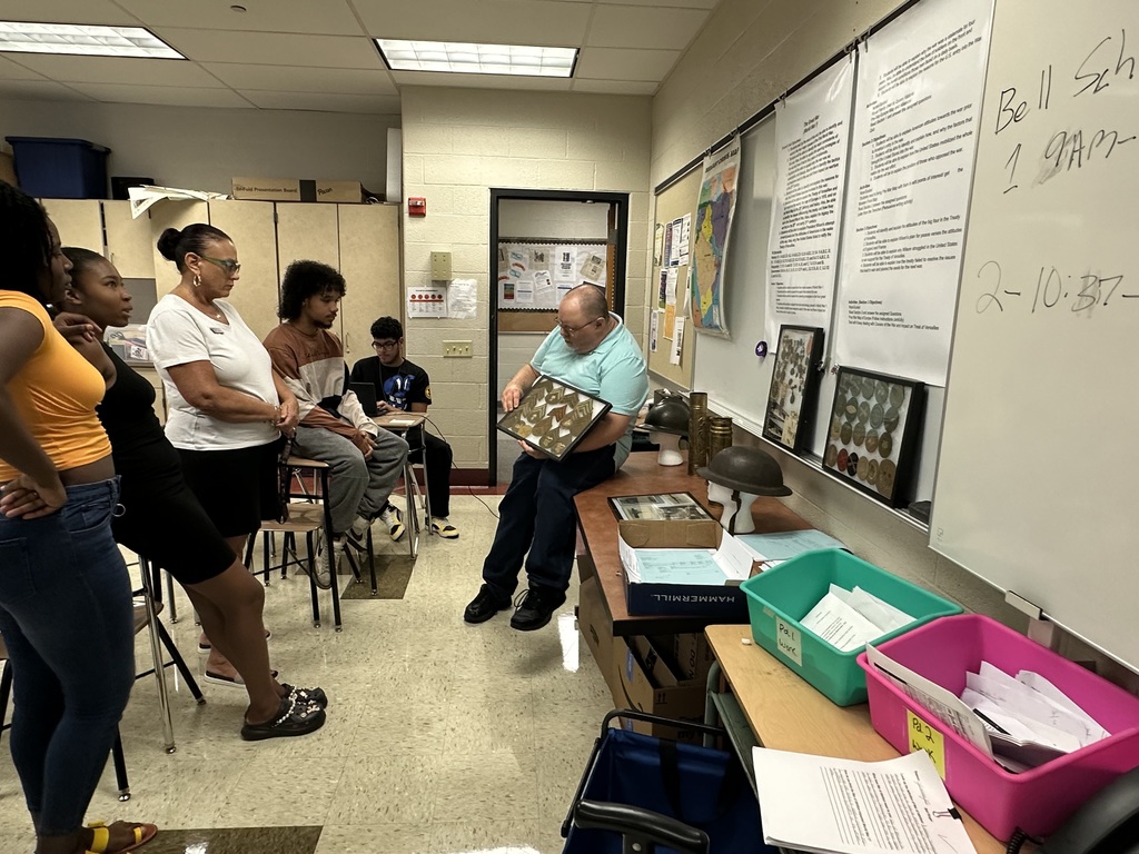 A group of individuals listens intently as a person demonstrates artifacts in a classroom filled with educational materials.