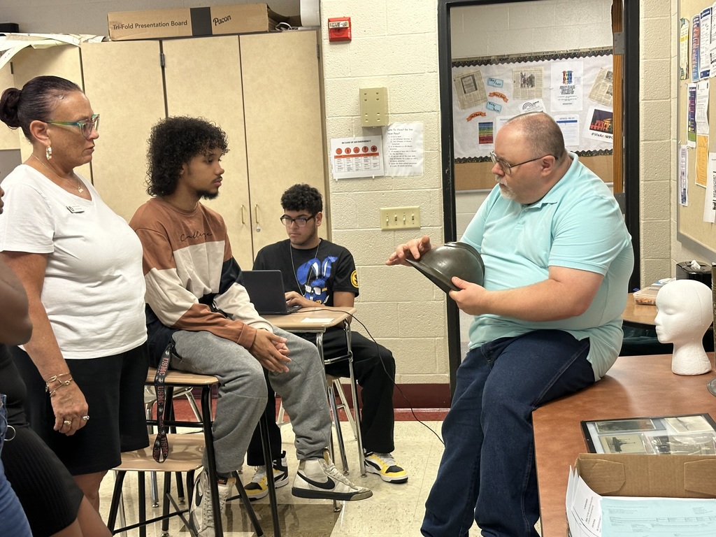 A teacher demonstrates an object to a group of seated students in a classroom setting, with materials displayed on a table.